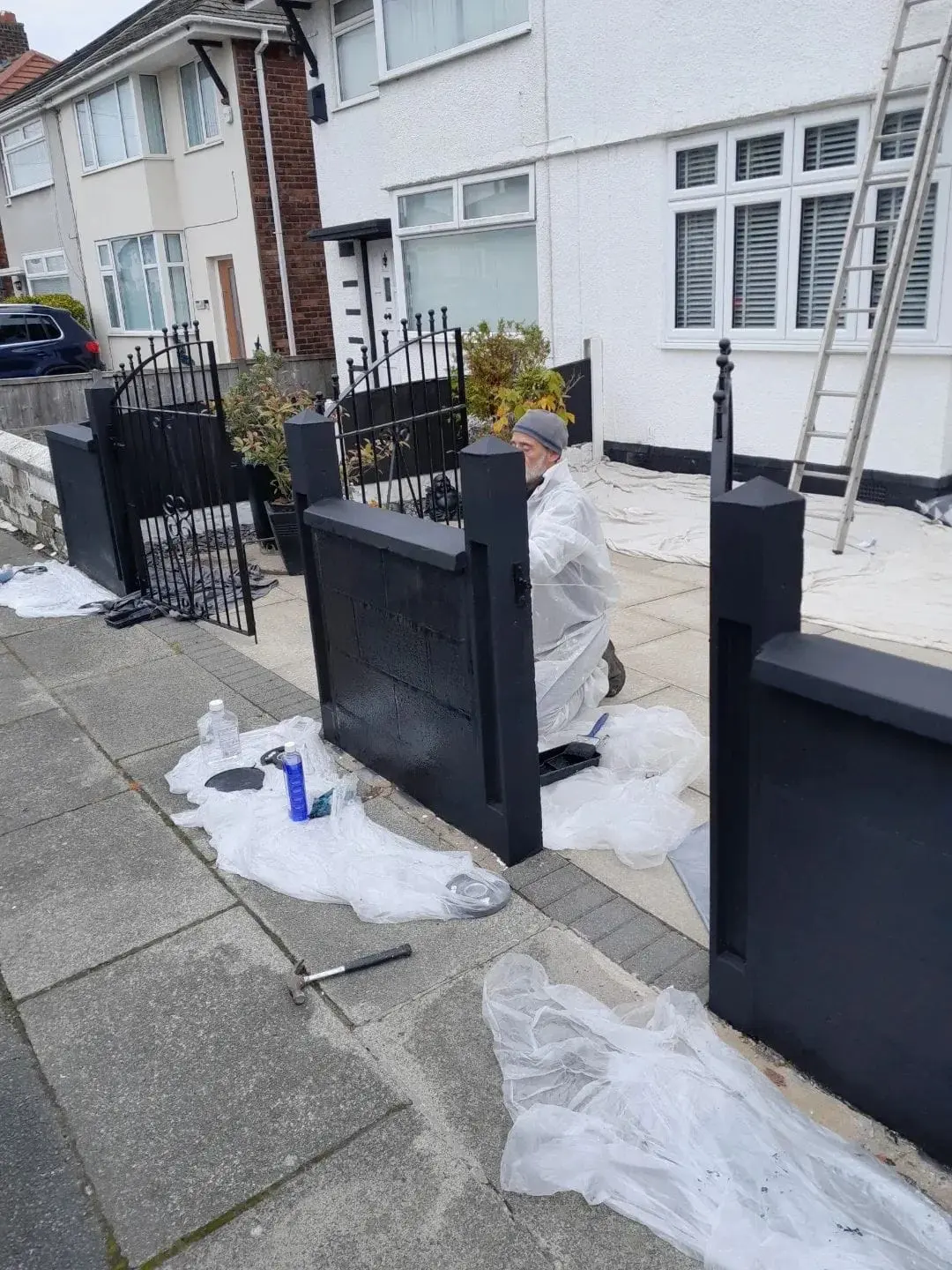 Painter working on a black front garden wall, with protective sheets on the ground and a ladder against a white house